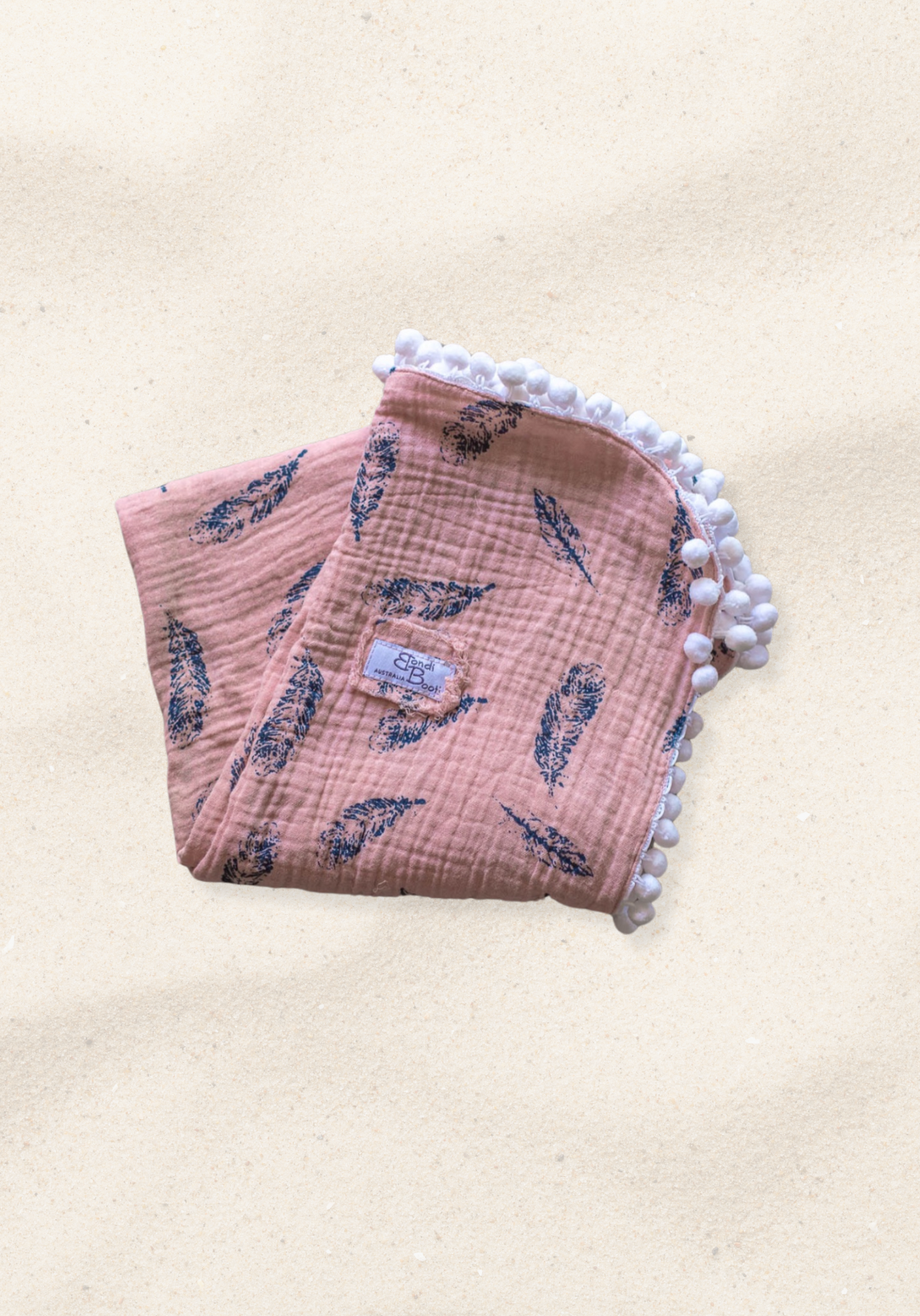 Dusty pink organic cotton baby blanket with navy feather print and white pom-pom trim, folded neatly with a visible Bondi Booti label.