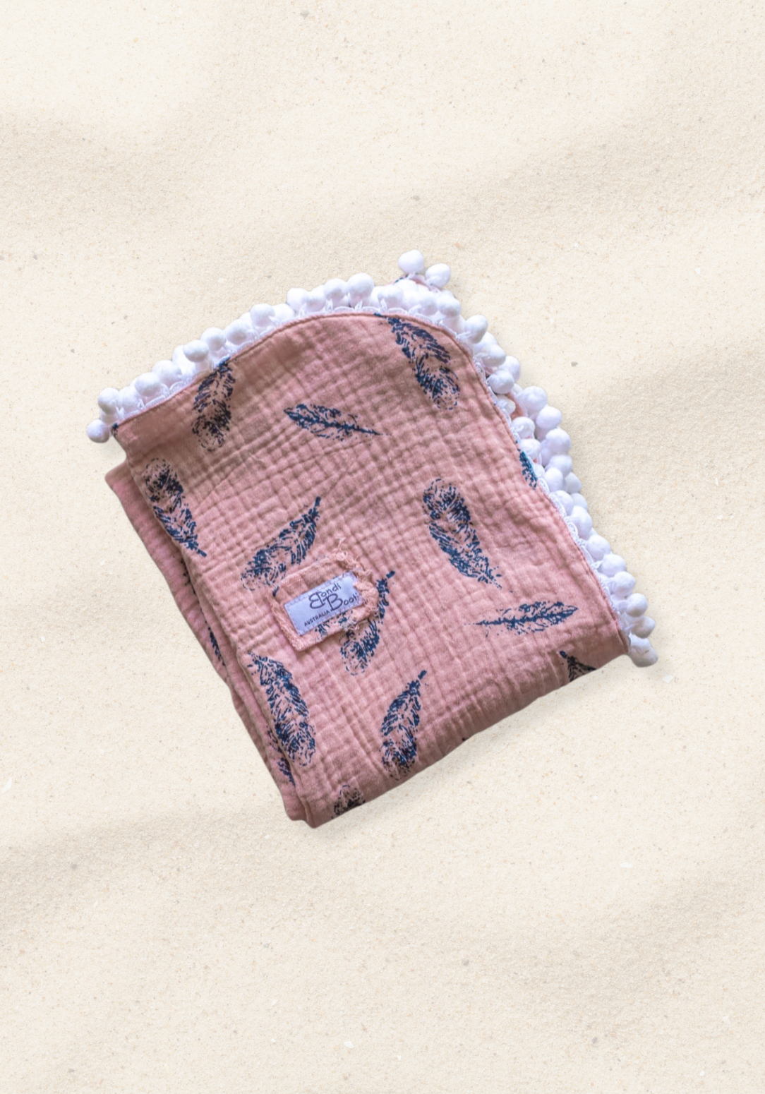 Dusty pink organic cotton baby blanket with navy feather print and white pom-pom trim, folded neatly with a visible Bondi Booti label.