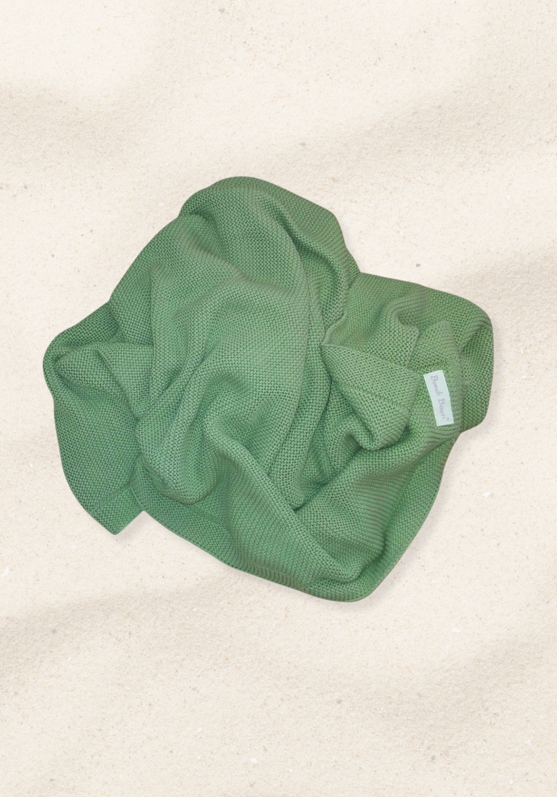 Folded green knitted baby blanket with a “Bondi Booti” label, placed on a light sandy background.