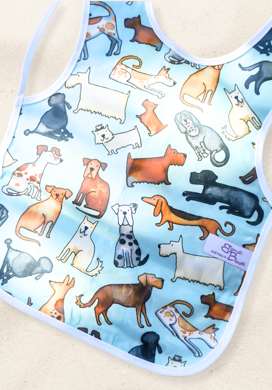 Laminated baby feeding bib featuring a playful pattern of illustrated dogs in various breeds and colors on a light blue background, trimmed in white with a snap closure, laid flat on a sand background.