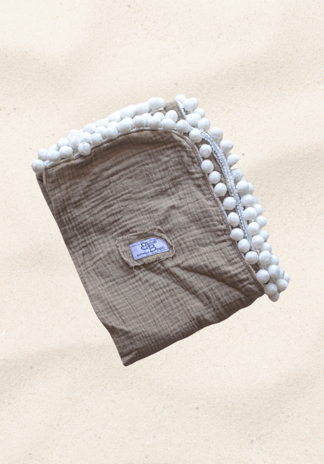 Taupe organic cotton baby blanket with white pom-pom trim, neatly folded with visible Bondi Booti label, displayed on a neutral background.