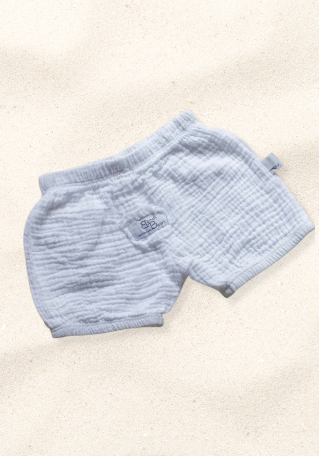 White organic cotton baby bloomers with soft elastic waist and leg openings, laid flat on a sand-colored background