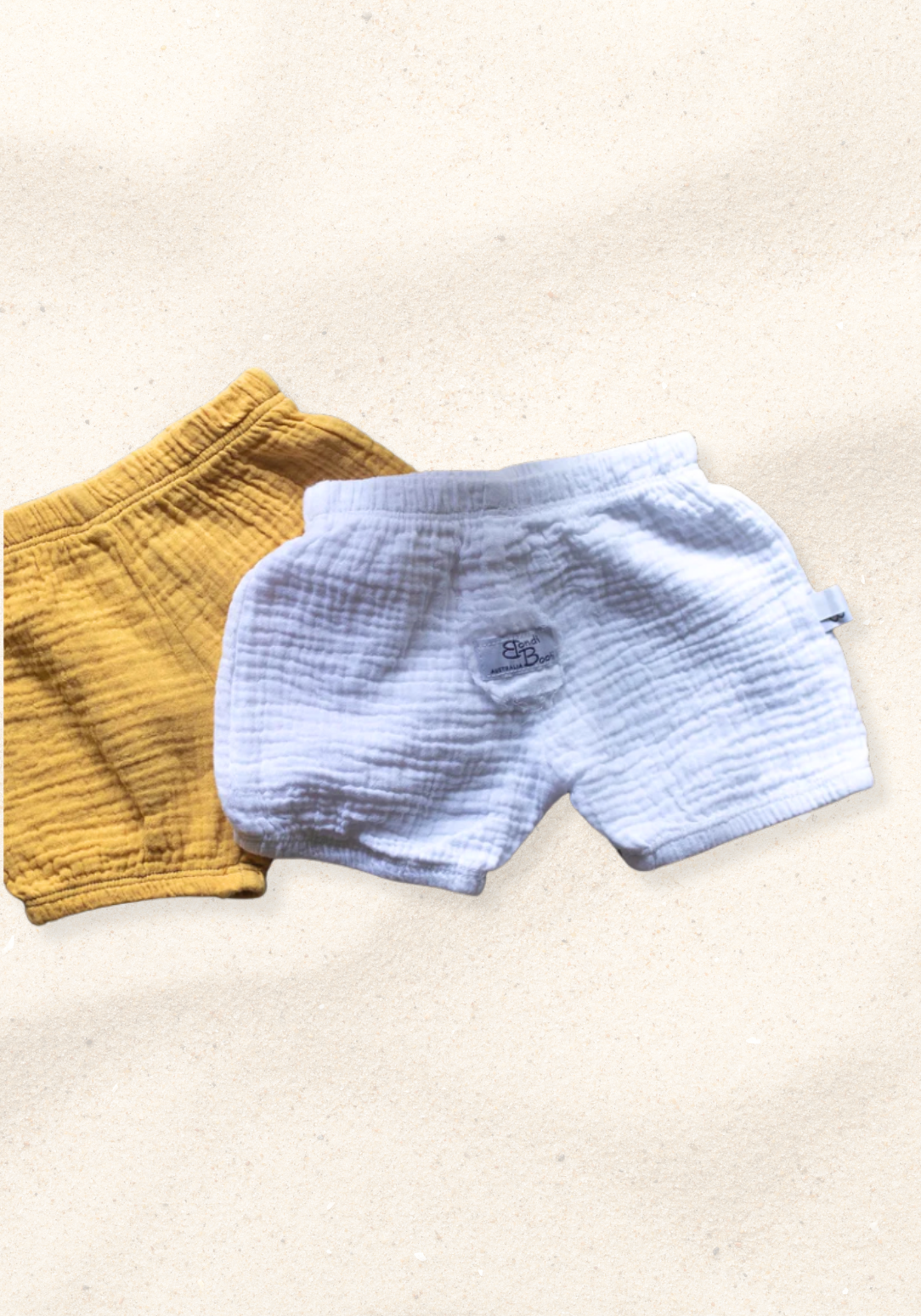 White organic cotton baby bloomers with soft elastic waist and leg openings, laid flat on a sand-colored background