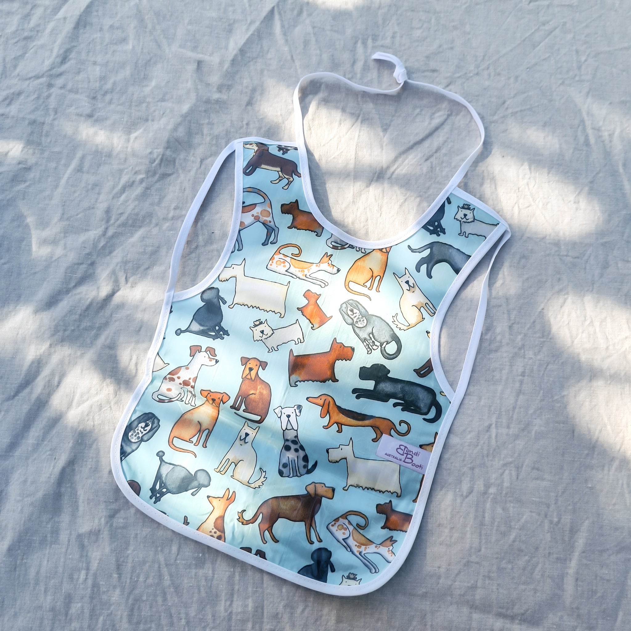 Laminated baby feeding bib featuring a playful pattern of illustrated dogs in various breeds and colors on a light blue background, trimmed in white with a snap closure, laid flat on a neutral fabric backdrop in soft natural light.