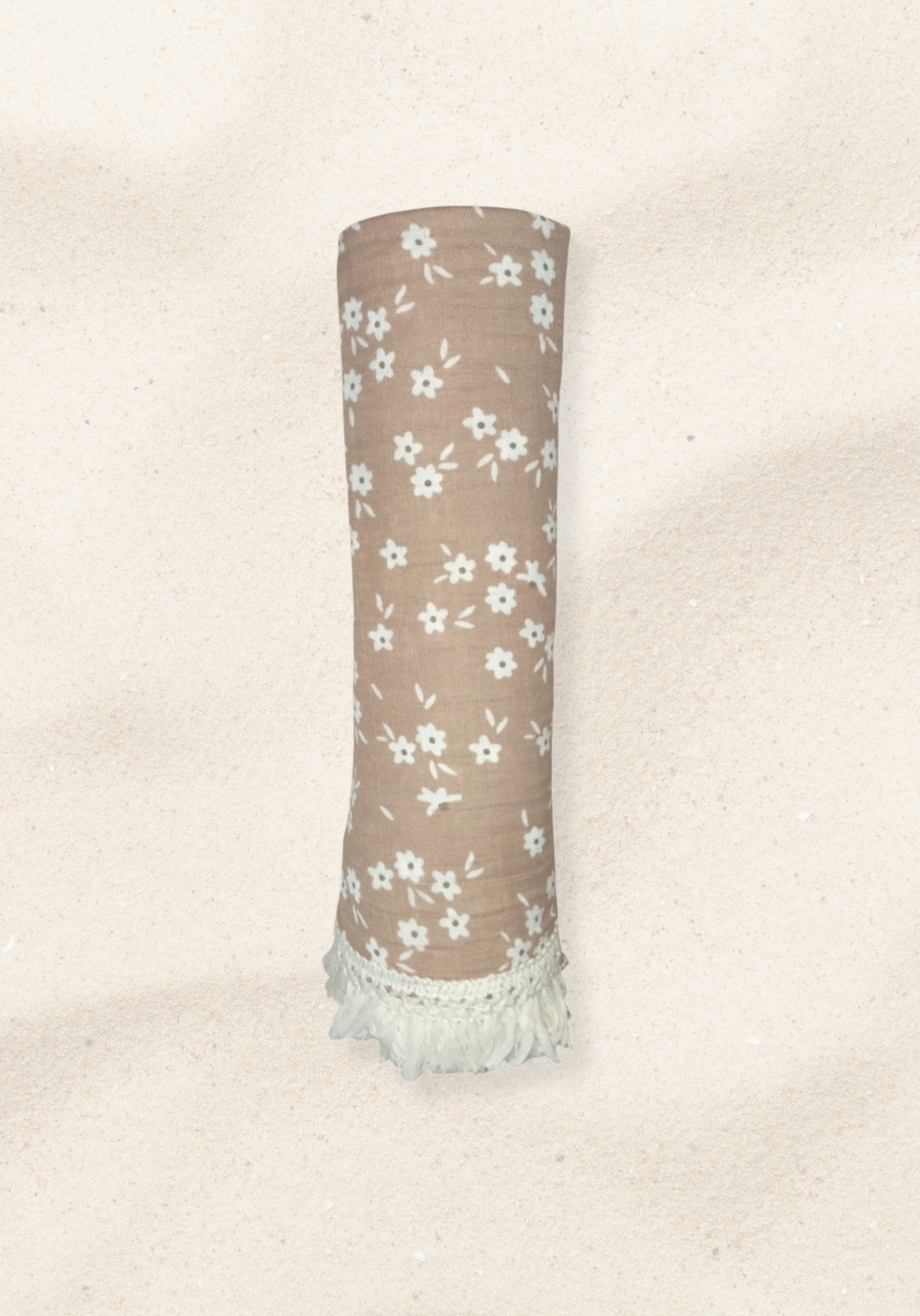 Rolled brown cotton baby blanket with white floral print and white fringed edges, placed on a light sandy background.