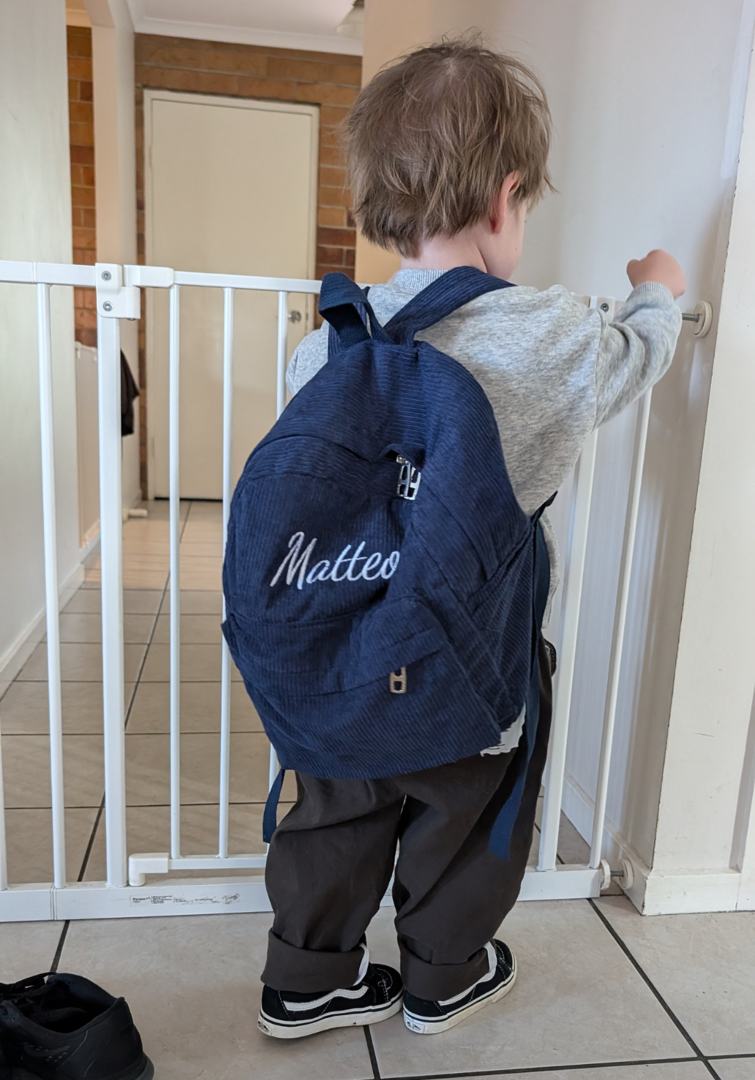 Toddler wearing navy blue personalized backpack with ‘Matteo’ embroidered, standing indoors near a baby safety gate