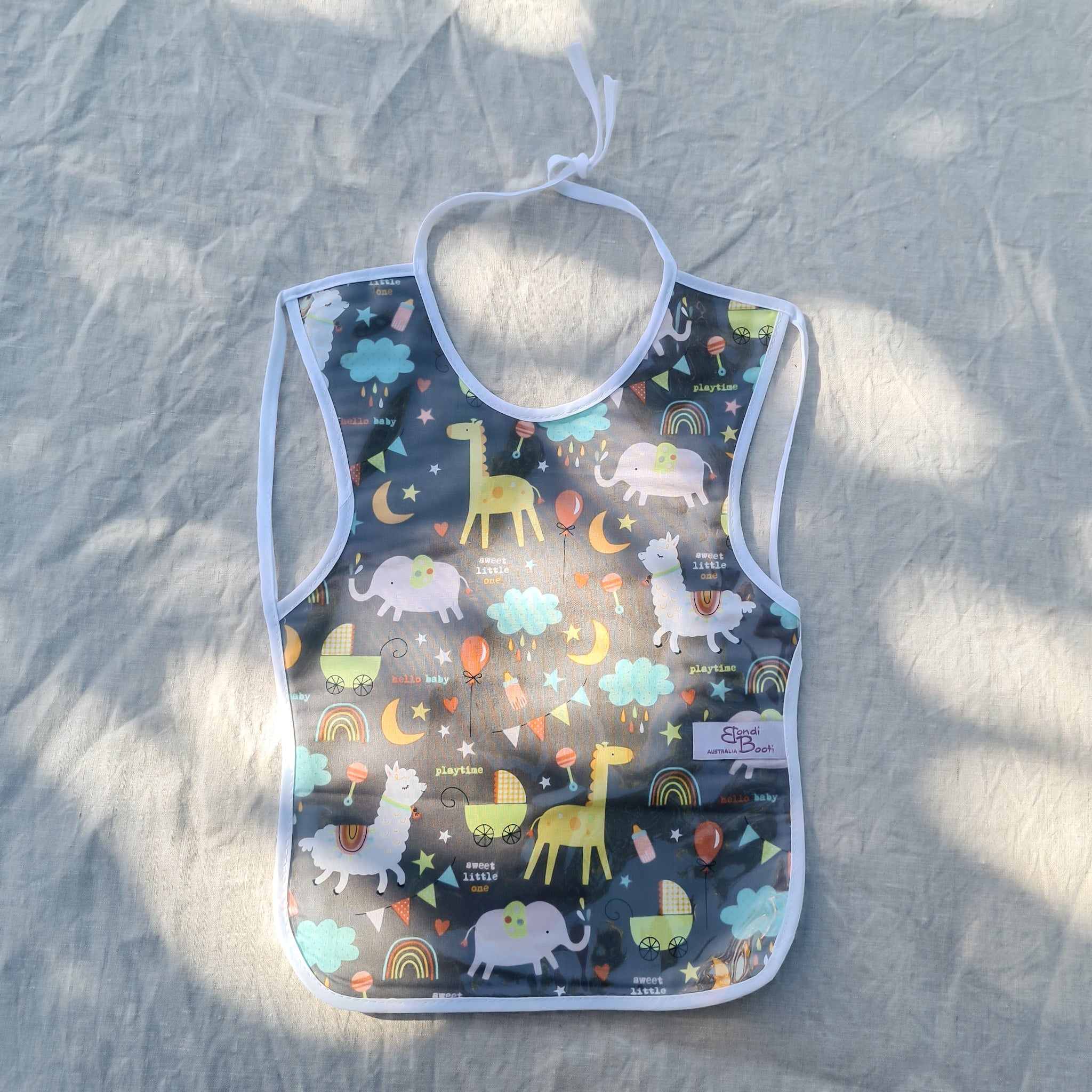 Laminated toddler feeding bib with a whimsical nighttime animal design, featuring giraffes, elephants, rainbows, and clouds on a dark grey background, with white trim and tie closure, laid flat on a light fabric surface under dappled light.