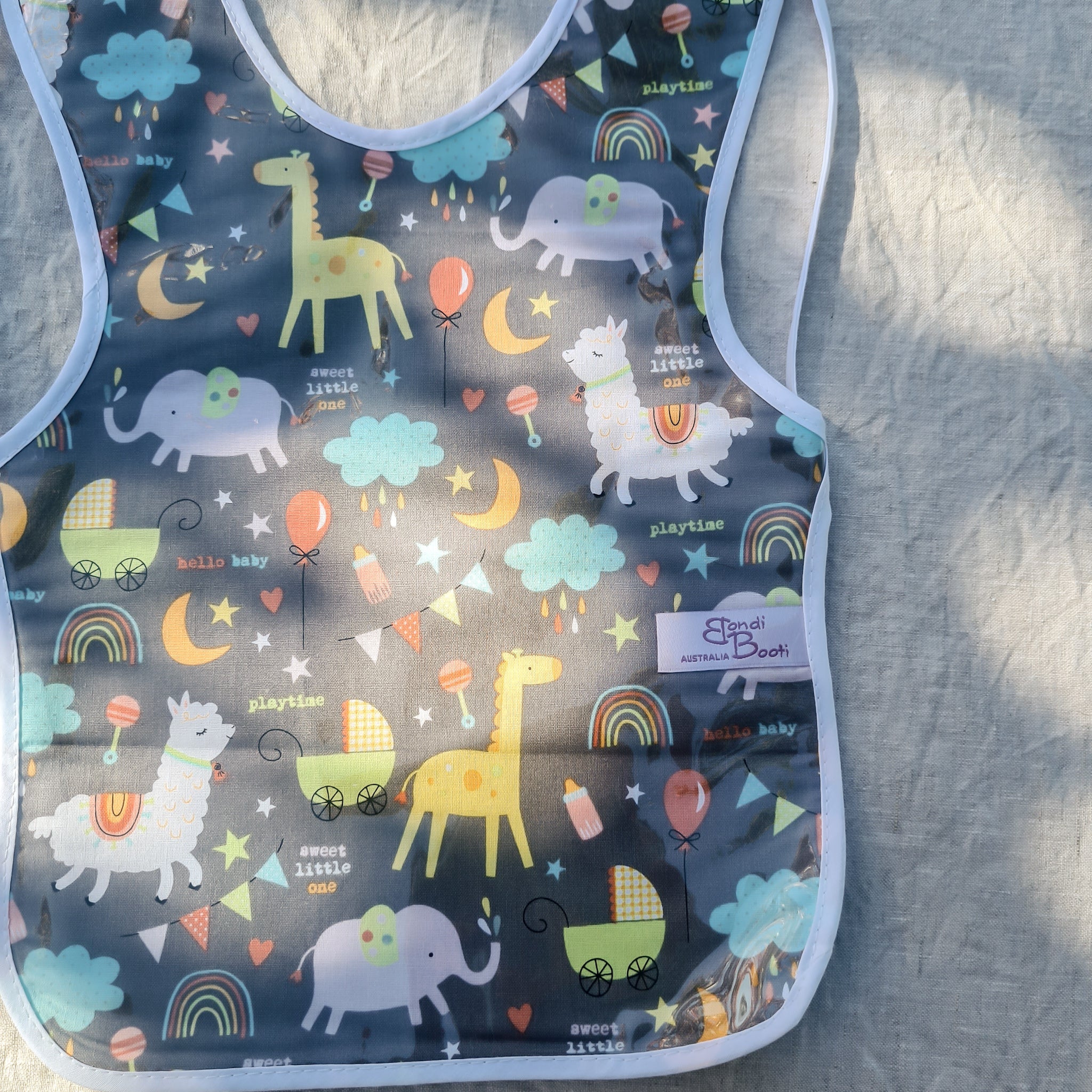 Laminated toddler feeding bib with a whimsical nighttime animal design, featuring giraffes, elephants, rainbows, and clouds on a dark grey background, with white trim and tie closure, laid flat on a light fabric surface under dappled light.